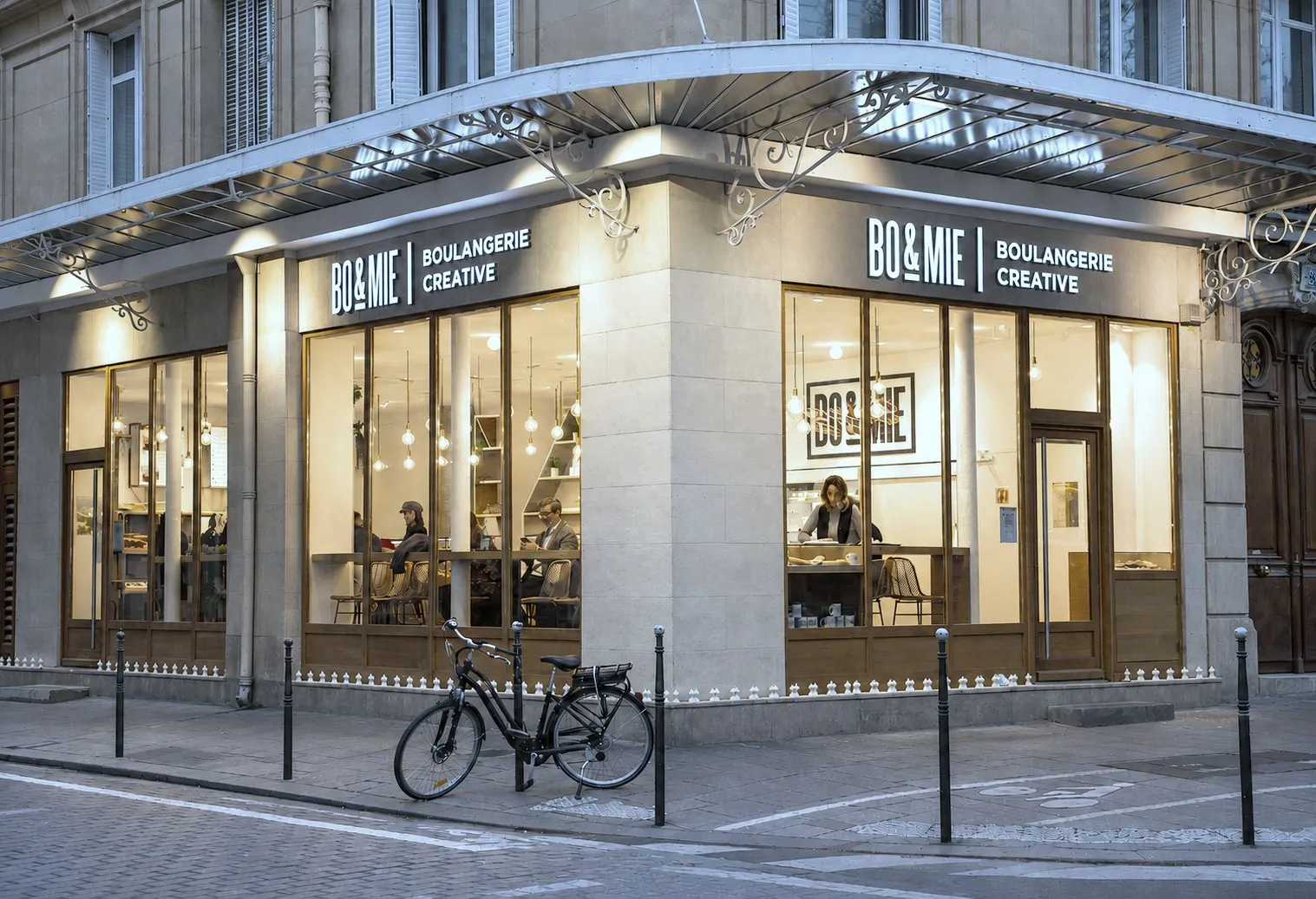 La boutique BO&MIE Saint-Martin au crépuscule : devanture d’angle éclairée, enseignes « Boulangerie créative » et clients en salle