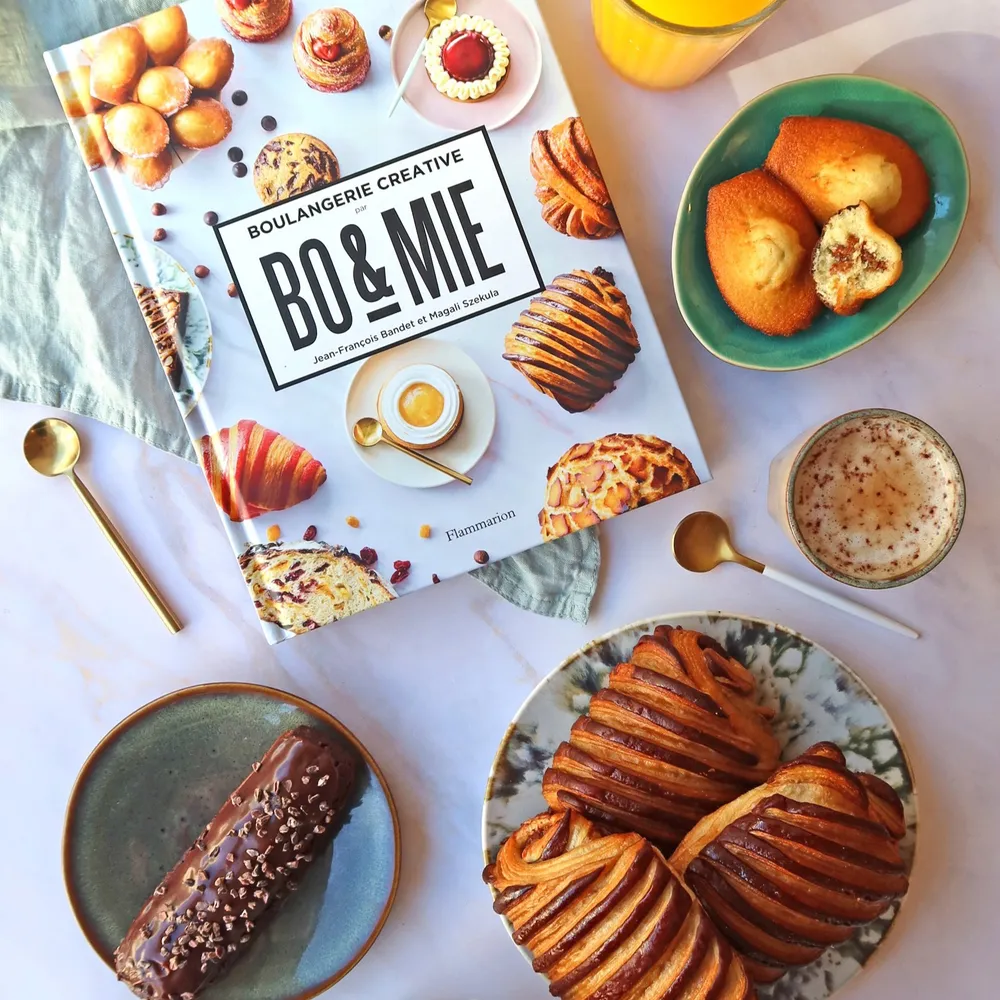 Le livre « Boulangerie créative par BO&MIE », chez Flammarion, posé sur une table de petit-déjeuner avec viennoiseries, madeleines et café